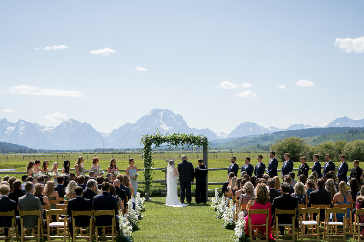 Bride and groom stand beneath a rustic wooden arch adorned with lush white and blue florals, set against the breathtaking backdrop of the Teton mountains.