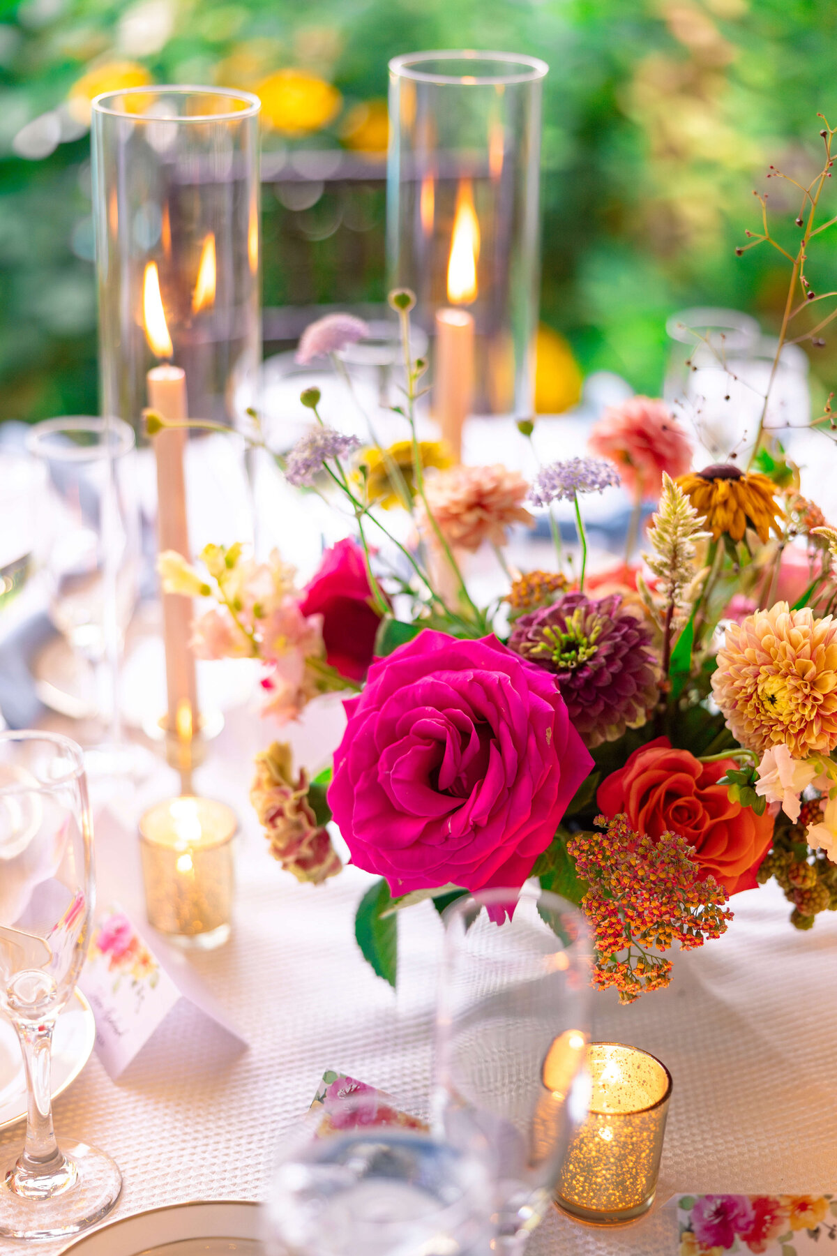 A beautifully set table features a vibrant floral arrangement with a prominent pink rose, surrounded by candles in glass holders. Soft lighting adds a warm, inviting ambiance to the elegant outdoor setting.