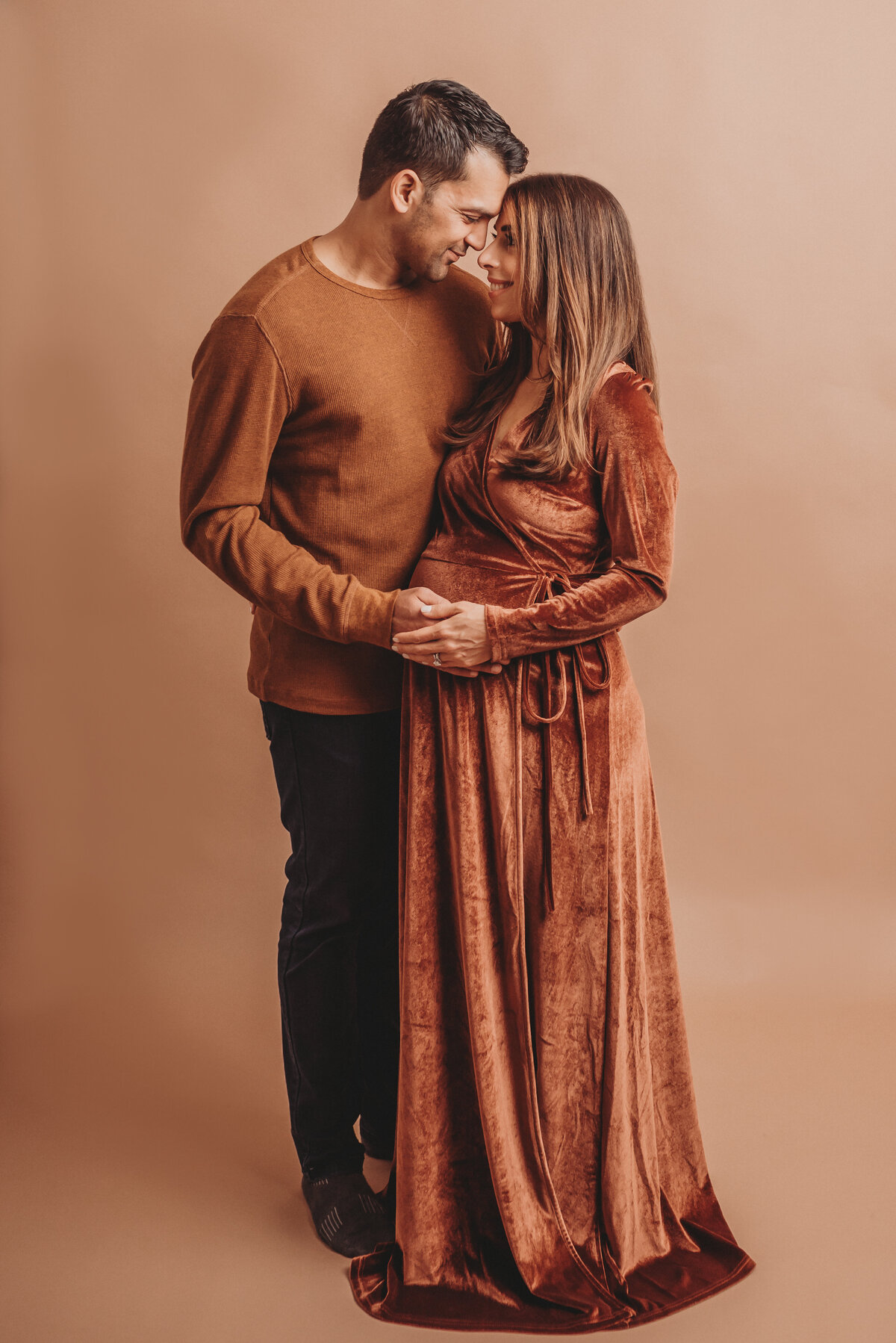 Couple at maternity photo shoot woman is wearing copper color floor length wrap dress and man is wearing brown long sleeve shirt with black slacks