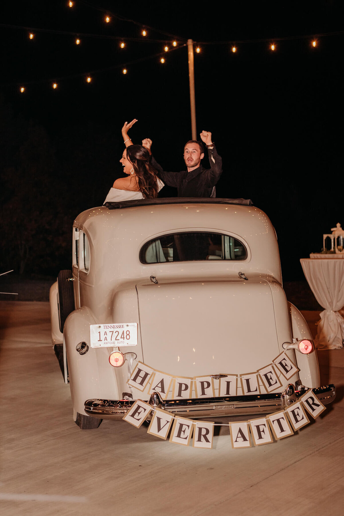 Photo of a bride and groom waving goodbye out of the rooftop of a white vintage vehicle with this sign happily ever after hanging off the back
