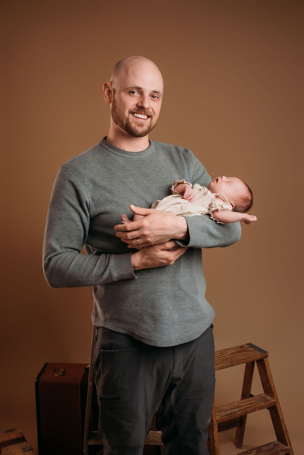 A man in gray stands in front of a neutral-toned backdrop holding a newborn baby in a beige outfit, captured by a photographer in Bemidji, Minnesota.