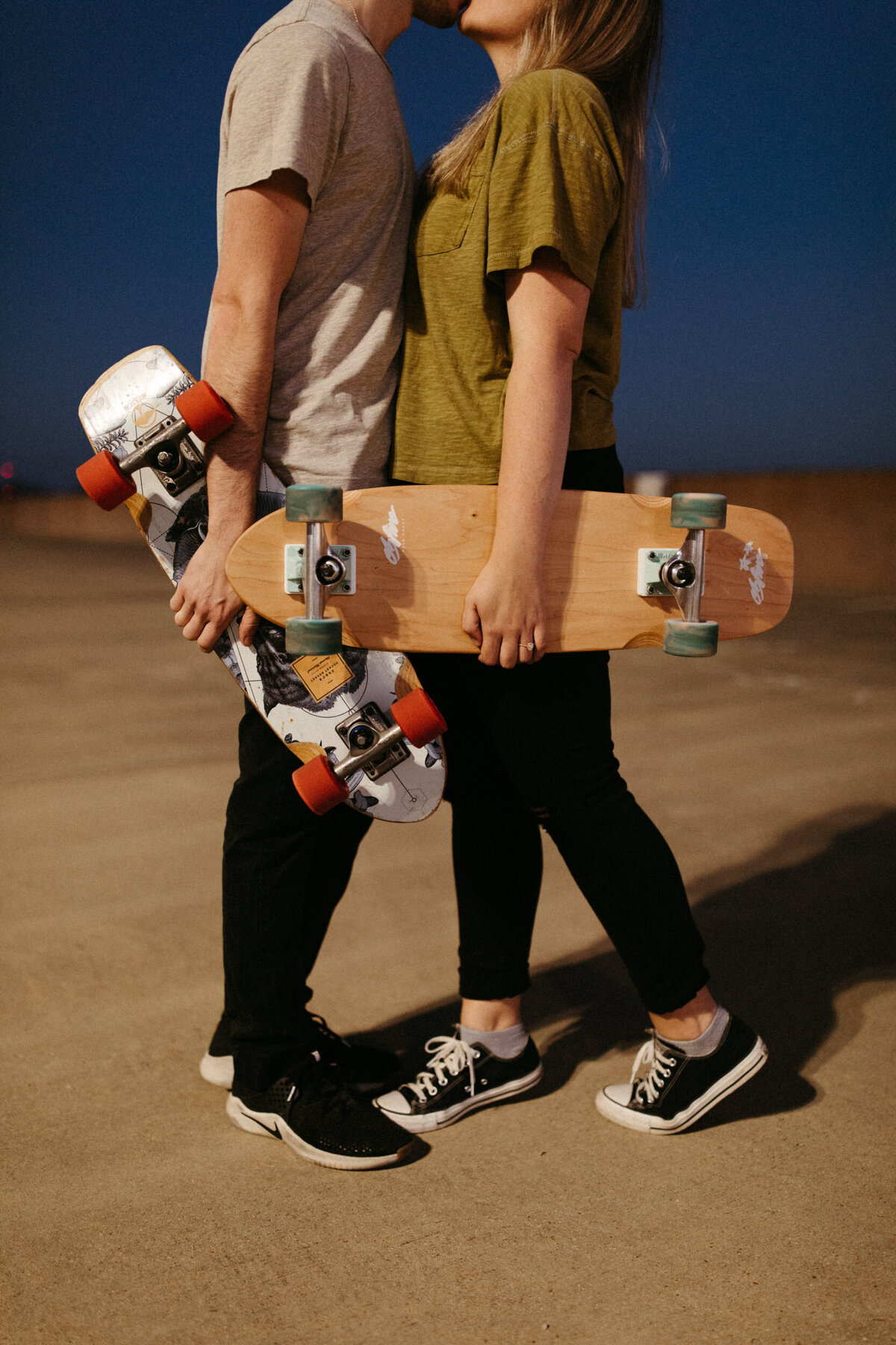 A couple is holding skateboards and kissing each other on top of a parking garage at night.