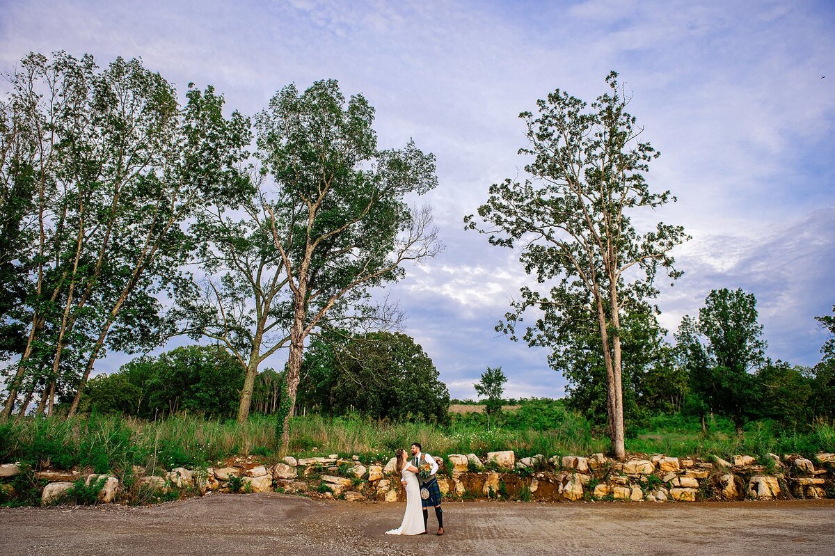 The bride, wearing a fitted wedding dress with a short train kisses the groom who is wearing traditional Scottish attire including a kilt and sporran. Behind them is a long wall of limestone and a green field with tall cedar trees at their Nashville elopement.
