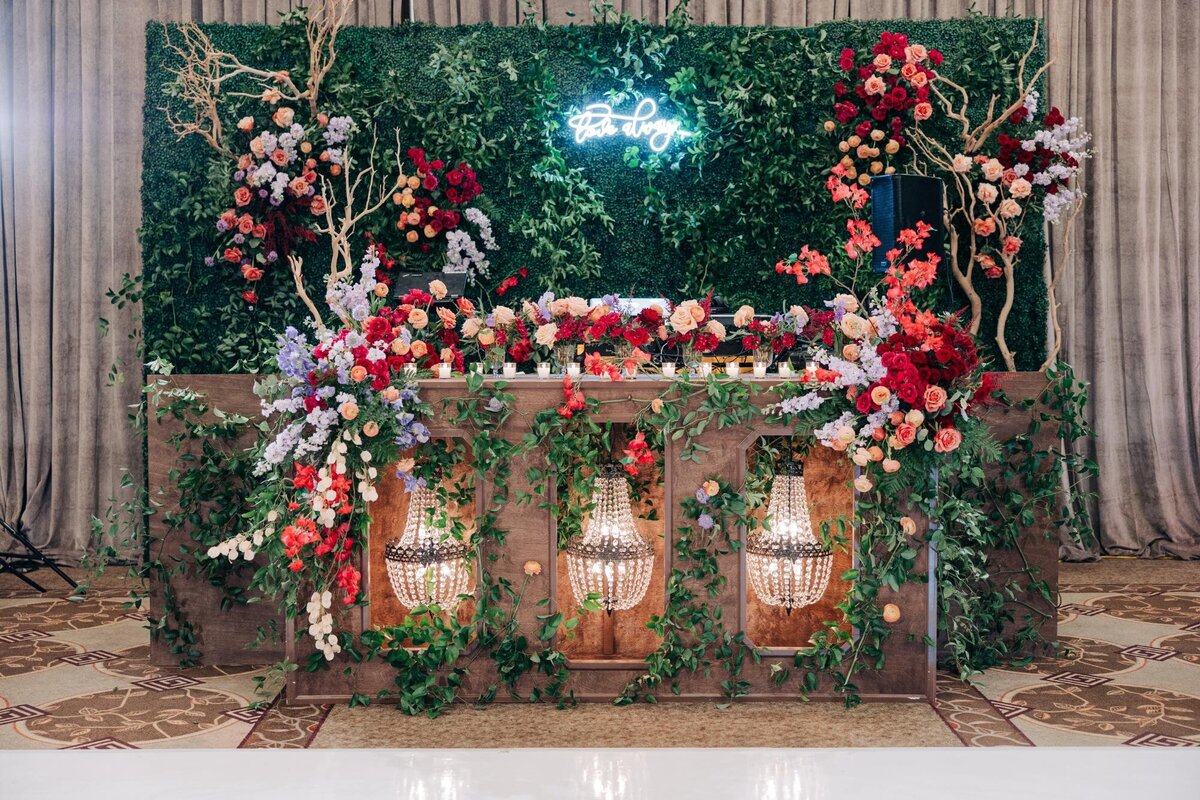 Elegant wedding reception table decorated with abundant flowers and greenery backdrop, and illuminated by two hanging lanterns.