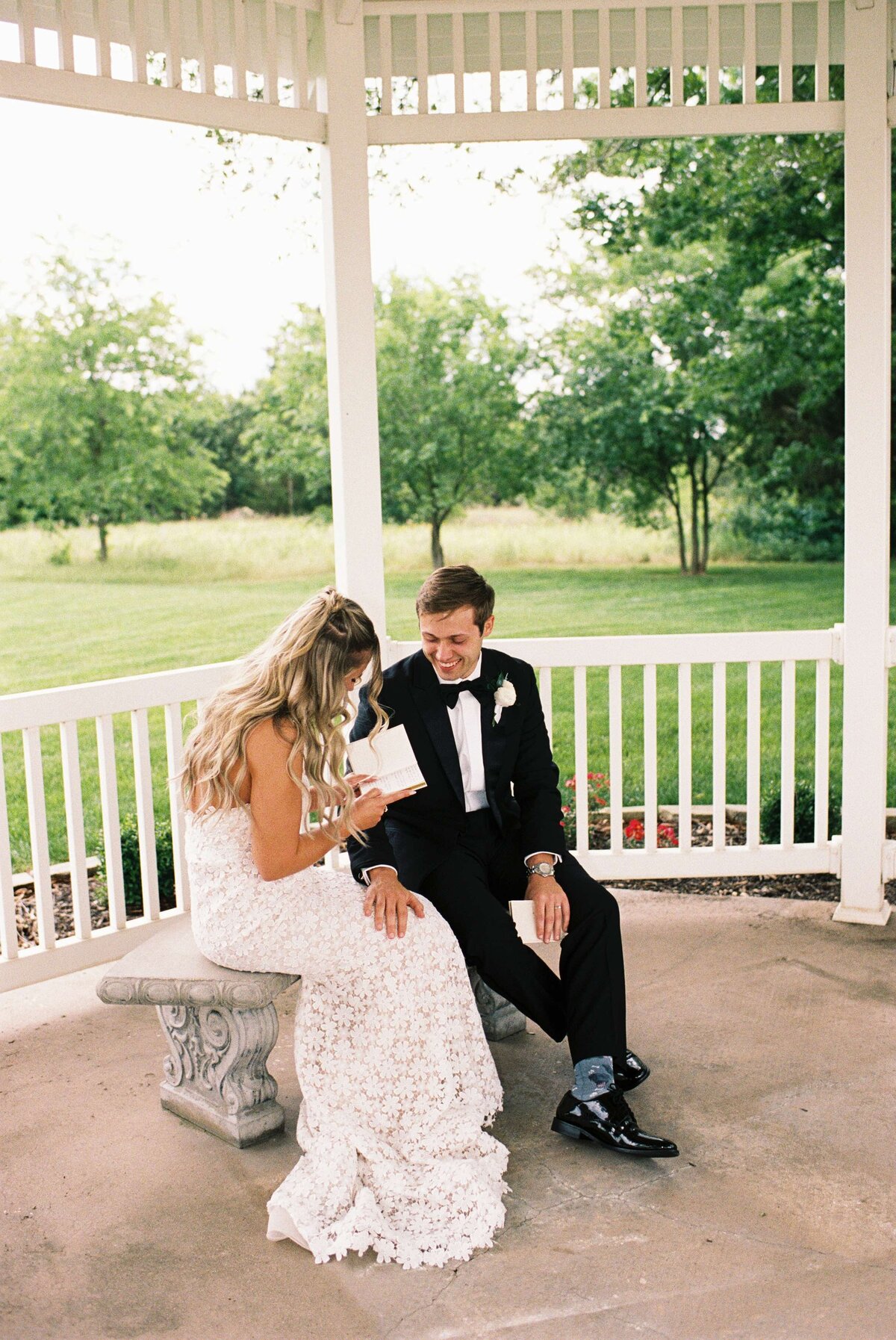 A bride in a white lace dress and a groom in a black tuxedo sit in a gazebo. The bride reads a card to the groom, sharing a tender, joyful moment.