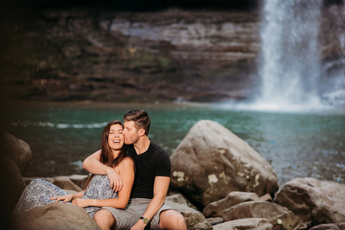 photo of a man kissing his fiancee's cheek in front of a waterfall