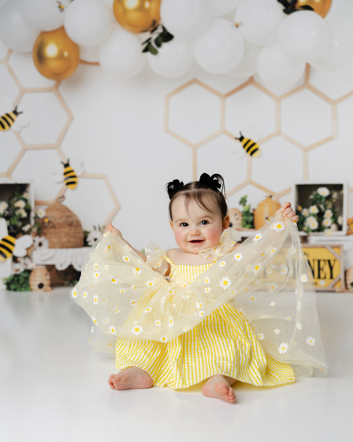 Smiling baby girl in a yellow gingham daisy dress holding up her sheer floral sleeves, seated in a bee-themed birthday setup with honeycomb accents and buzzing bees. Photograph by Yucaipa cake smash photographer Kaitlyn Dawn Photography.