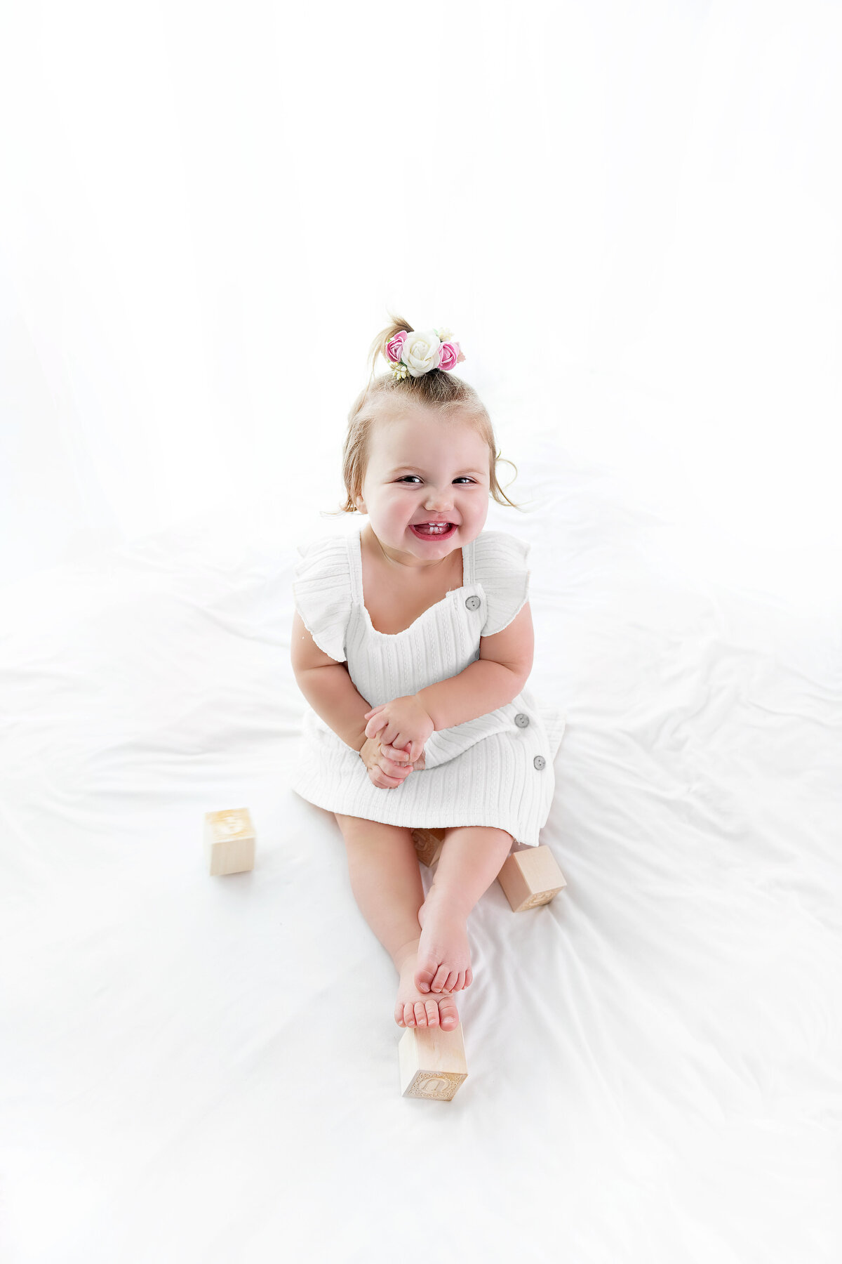 A toddler girl sits on a bed in a white dress smiling and playing with wooden blocks as posed by a childrens photographer Atlanta