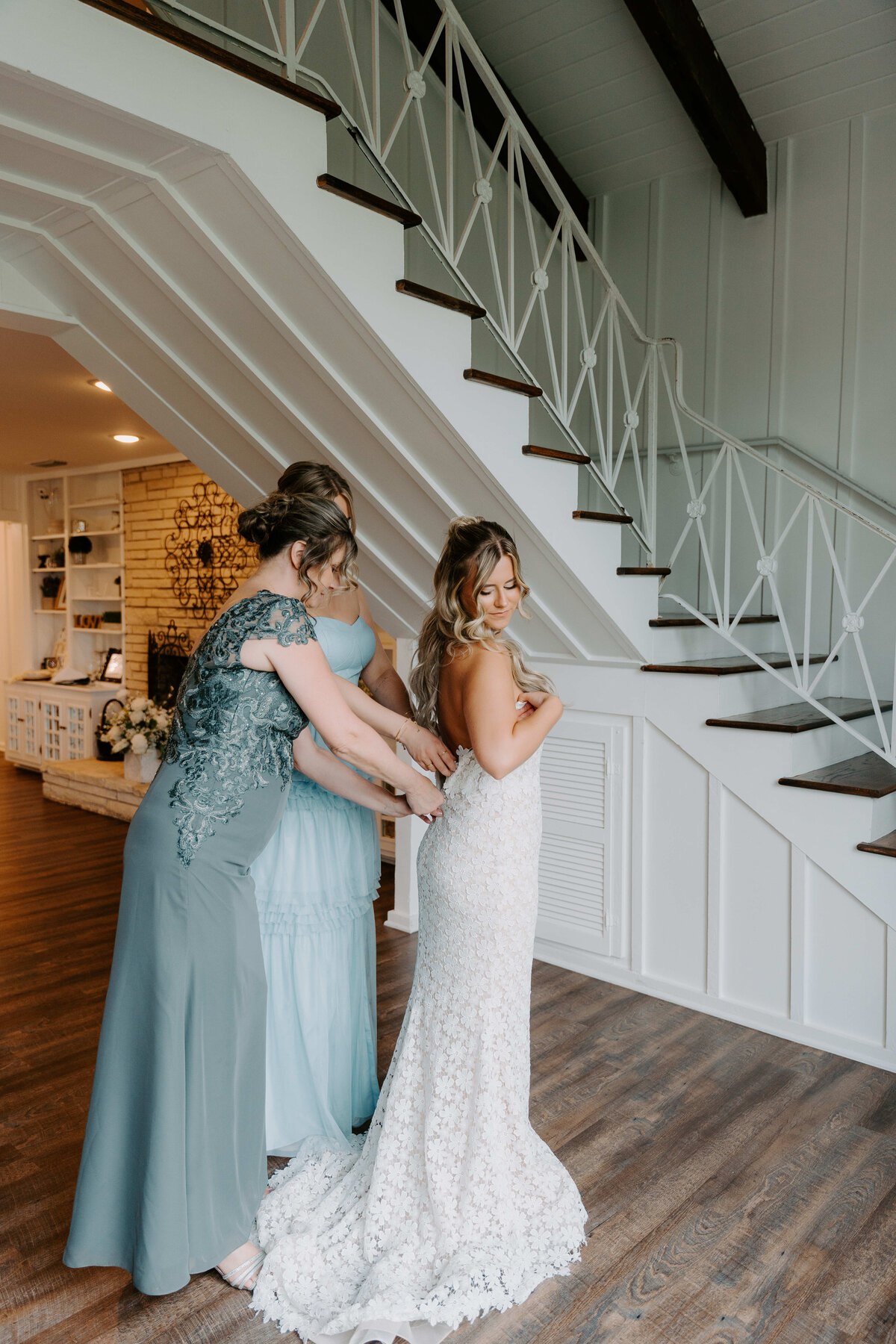 A bride in a lace gown is being assisted by two women in pastel dresses under a staircase. The atmosphere is calm and joyful.
