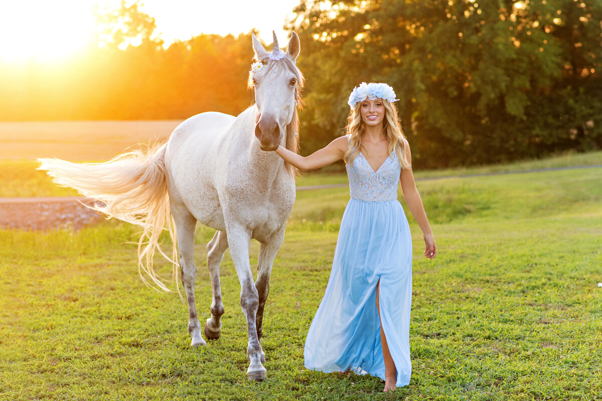 High school senior girl wearing long blue dress  and flower crown walks across field with her white horse.