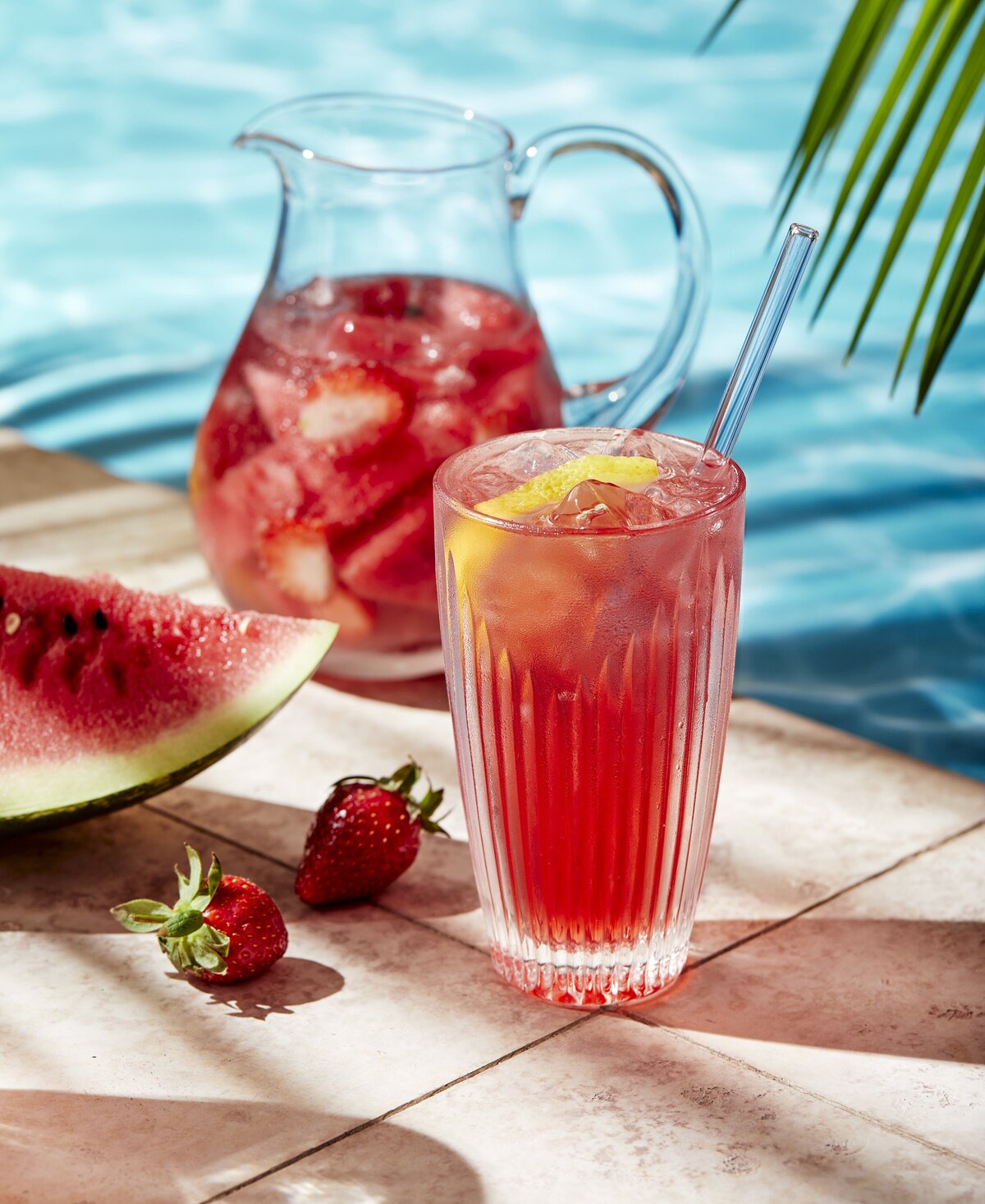 A fruity drink with a pitcher of it behind sitting next to a pool.