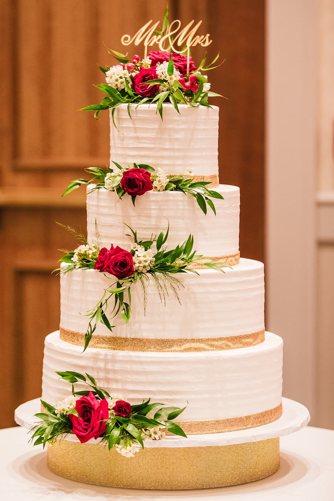 A multi-tiered wedding cake adorned with red roses and green foliage, topped with a 'Mr & Mrs' golden cake topper.