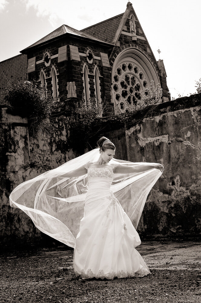 bride wearing a trumpet style wedding dress, holding a long veil blowing in the wind in front of the church