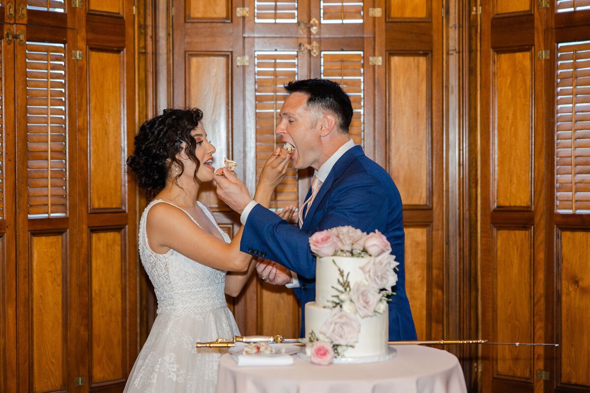 Bride and groom feeding each other cake in a room with wooden paneling, a table with a floral centerpiece, and a cake stand during an Iowa wedding.