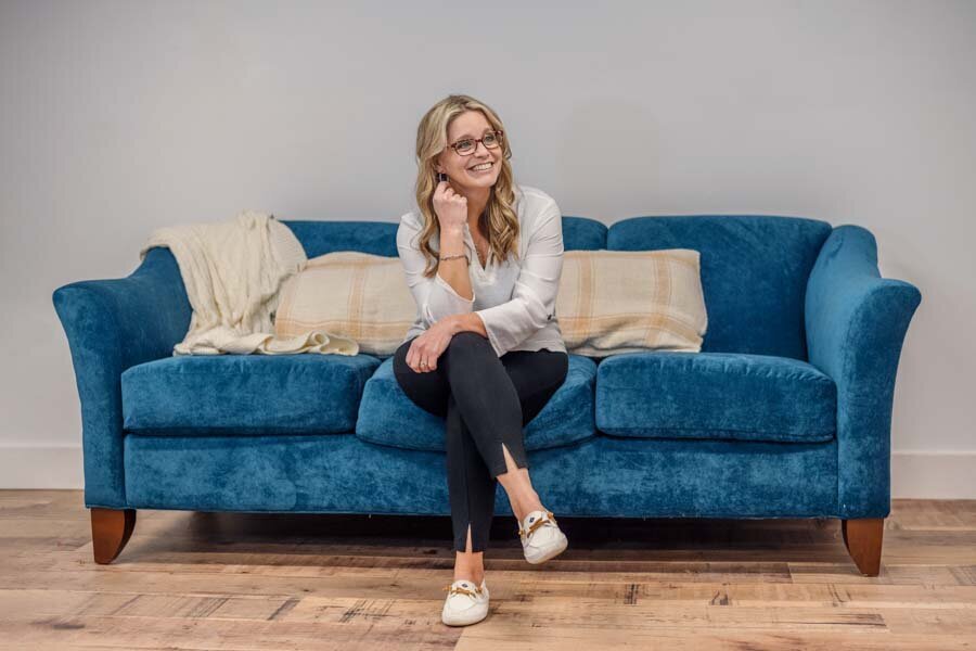 A woman in glasses, smiling and sitting on a blue sofa with a cream throw blanket. she wears a white blouse, black pants, and white shoes.