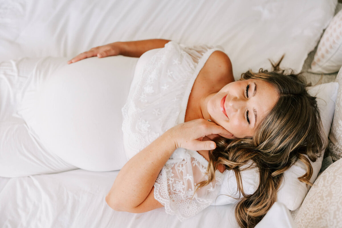 pregnant woman wearing white, laying on white bed with hand on cheek smiling