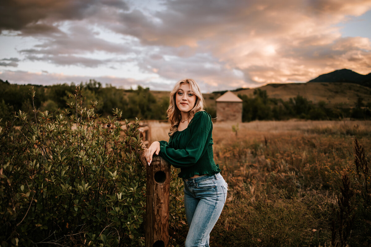 girl leans up against fence in boulder colorado for senior photos with cotton candy skies behind her