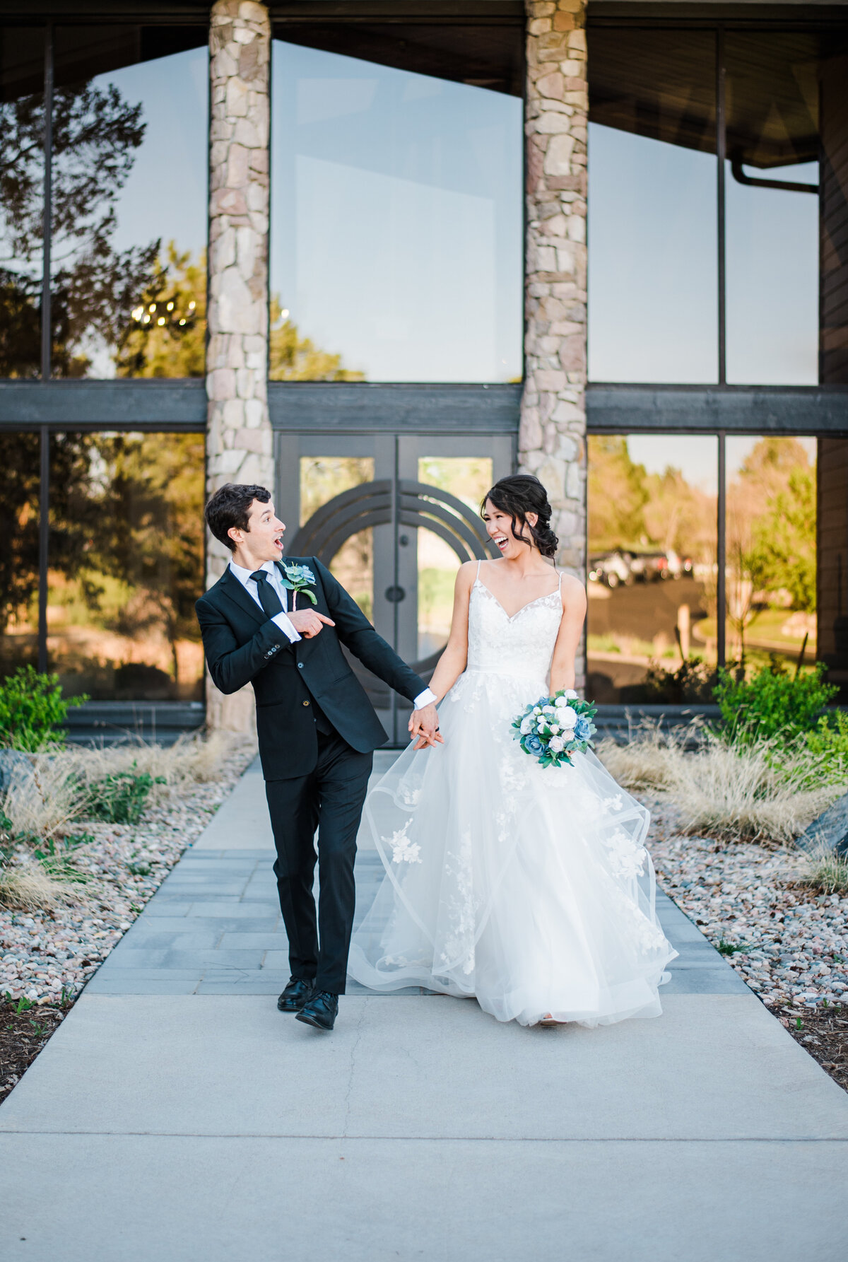 bride and groom holding hands and walking on a path at their denver wedding venue as the groom leans and points to his bride