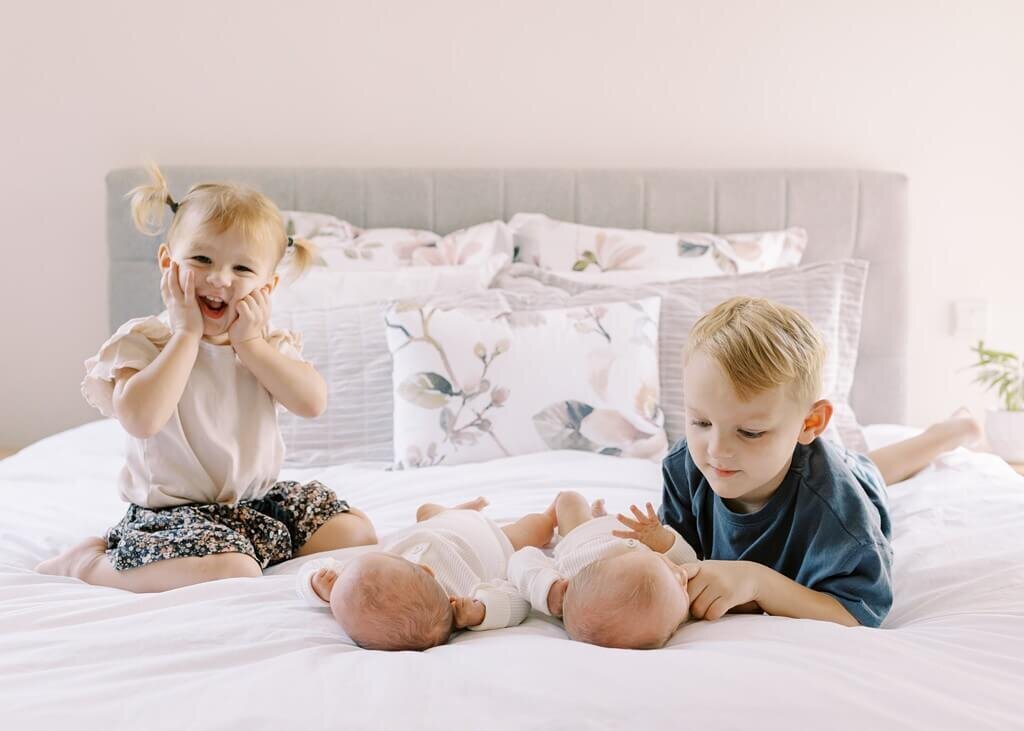Two older siblings lying on a bed gently touching their newborn twin siblings.