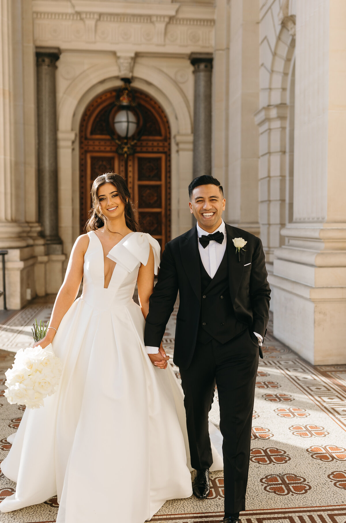 An image of a smiling bride and groom. They are walking hand in hand with the bride on the left and the groom on the right. The bride is holding a white bouquet in her  right hand and the groom has his left hand in his pocket/