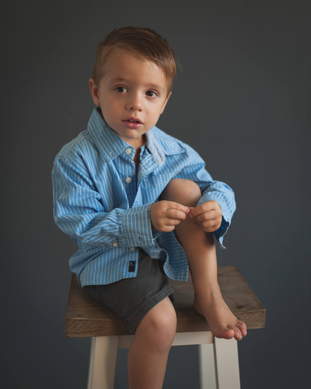 studio portrait of a preschool-aged boy on a stool  looking away wearing a blue  dress shirt and grey shorts, barefoot, with a grey backdrop