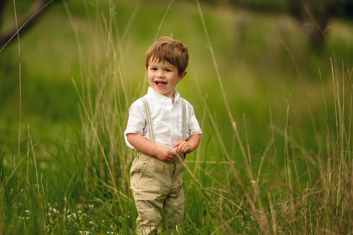 A toddler standing in a field of wildflowers gives a big smile while wearing khaki pants and suspenders for his portrait.
