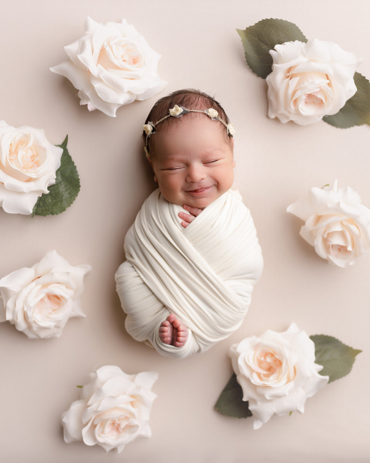 Newborn photoshoot in West Palm Beach Florida. Baby is wrapped in a knit swaddle with her toes and fingers peeking out of the wrap. Baby is wearing a delicate floral headband and surrounded by large rose blooms. Baby is sleeping and has a big smile.