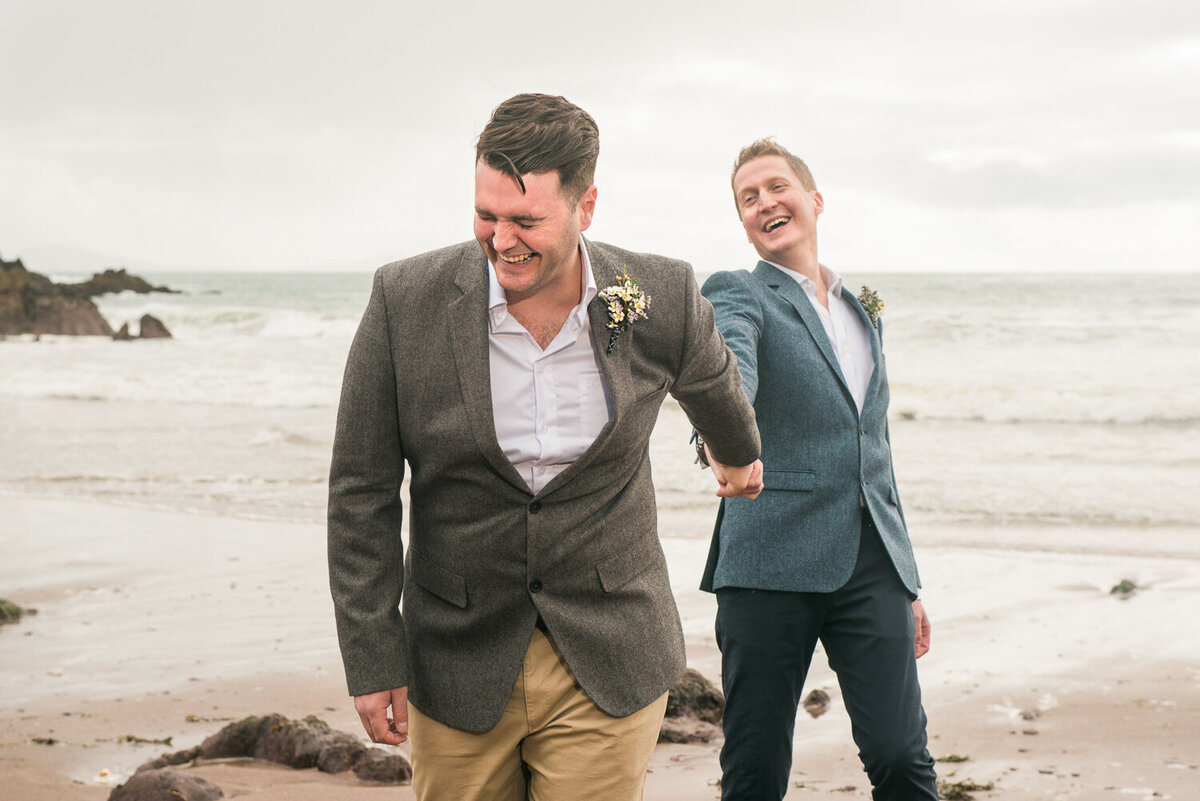 Gay male wedding couple holding hands and laughing on beach