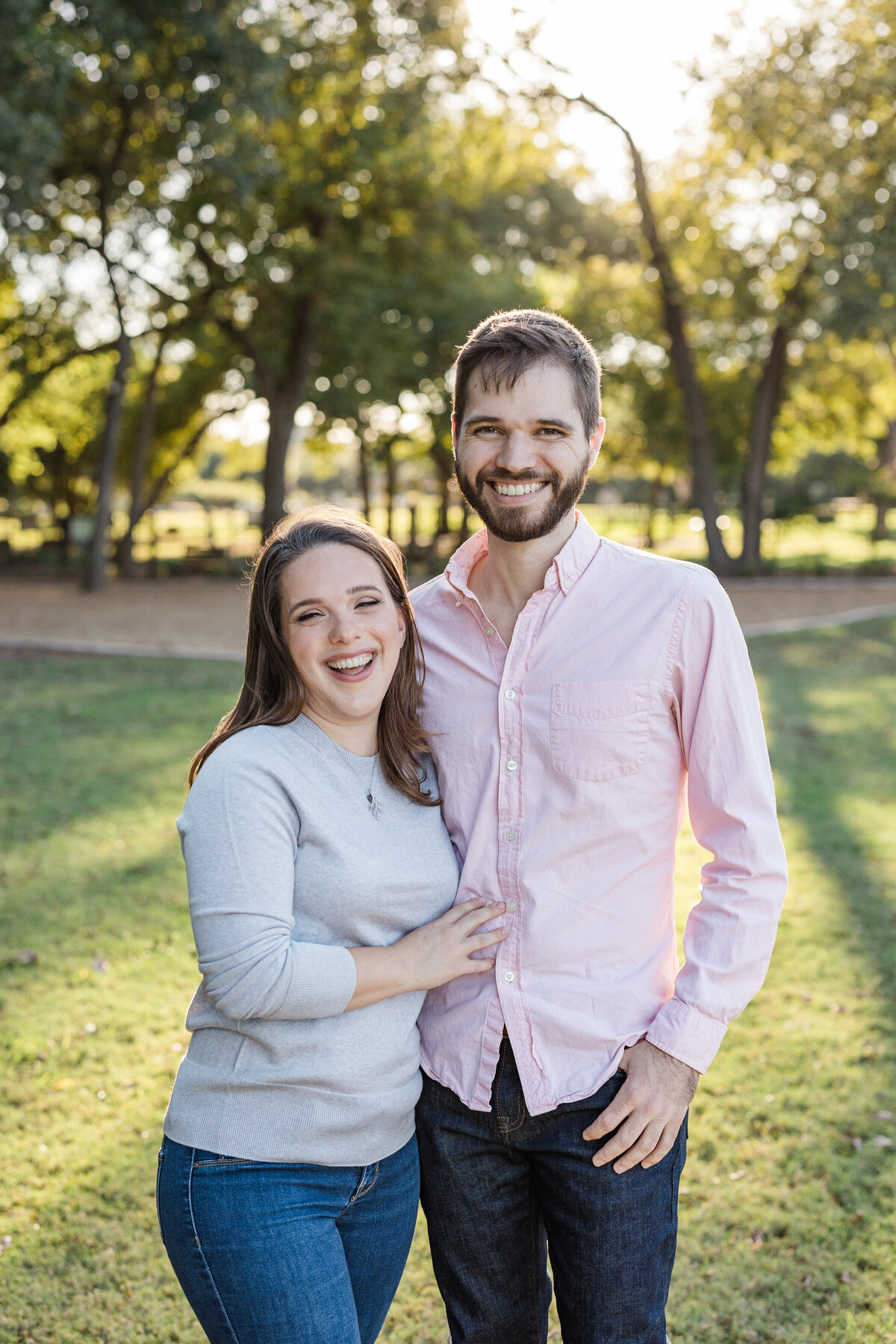 A couple posing joyfully together in a park during their engagement photoshoot in Dallas, Texas. The woman on the left is wearing a grey top, a necklace and jeans. The man on the right is wearing a salmon dress shirt and dark jeans.