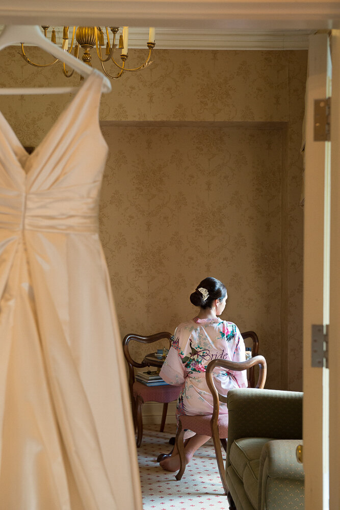 brunette bride wearing a headpiece and floral pink kimono style dressing gown in the bridal suite of Cahernane House with her wedding dress hanging over the door