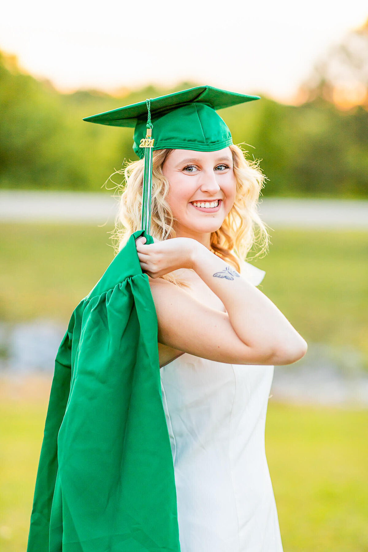 Senior graduate in a white dress holds her green cap and gown over her shoulder