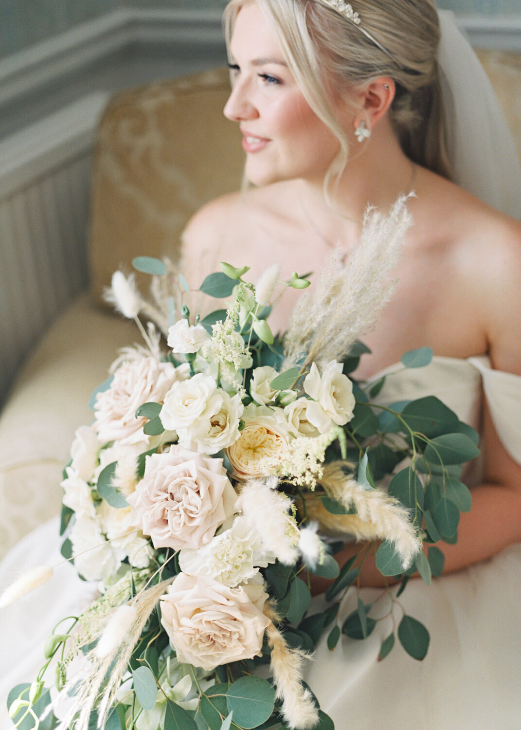 bride sitting looking out of window and and holding light blush and white cascade flower bouquet