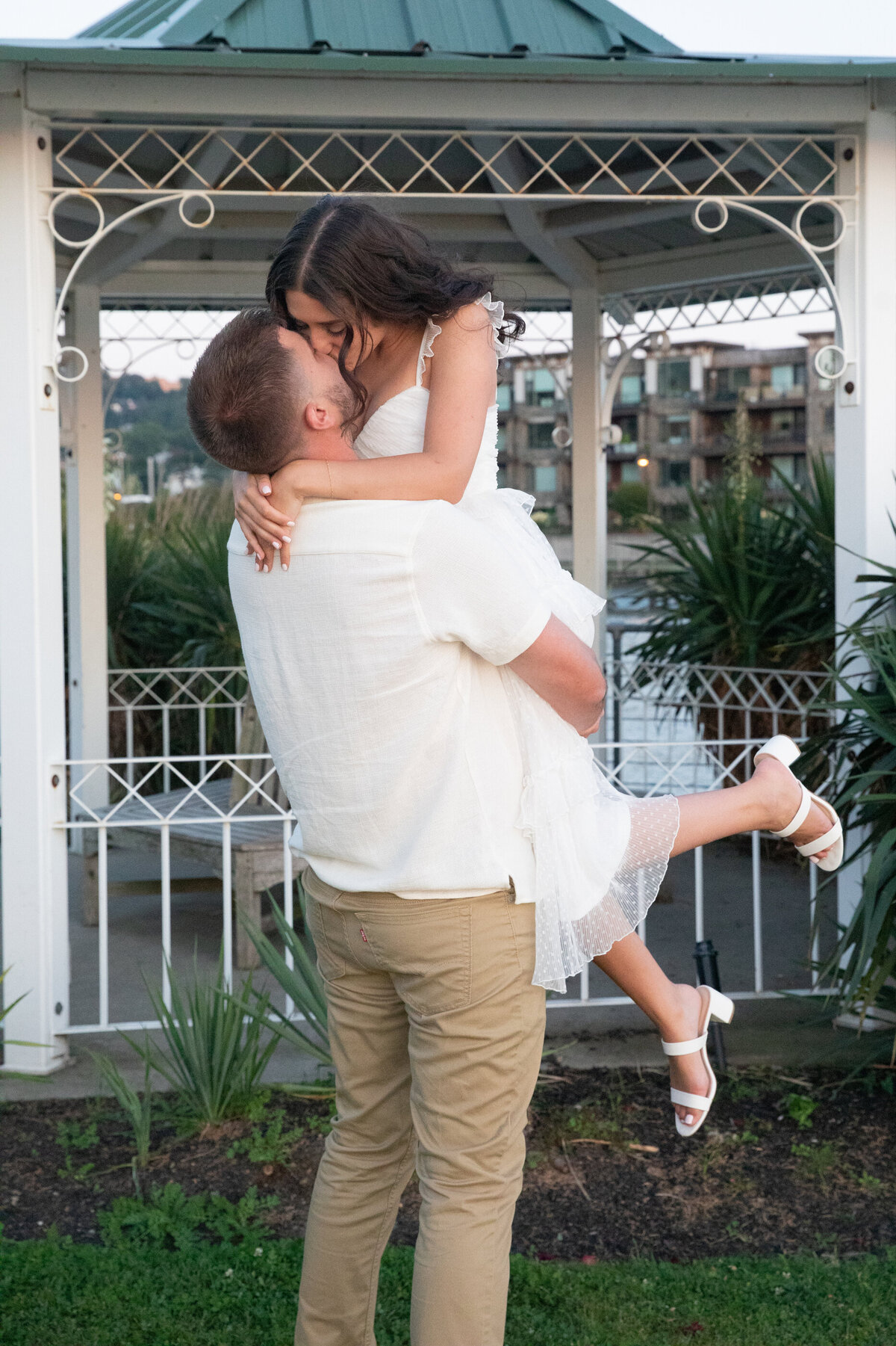 A man in a white shirt and khakis picks up his fiance in front of a gazebo engagement session New York wedding photographer