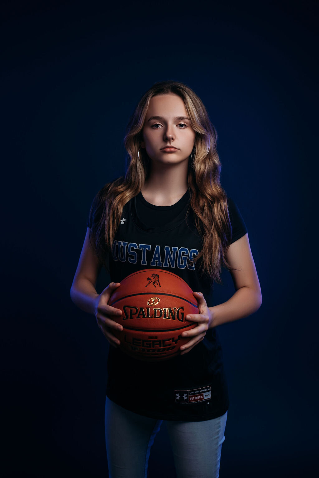 A teenage girl in a sports jersey holds a basketball in front of a blue solid backdrop illuminated by a blue glow around her for her senior graduation photoshoot, captured by a fine art studio photographer in Bemidji, Minnesota