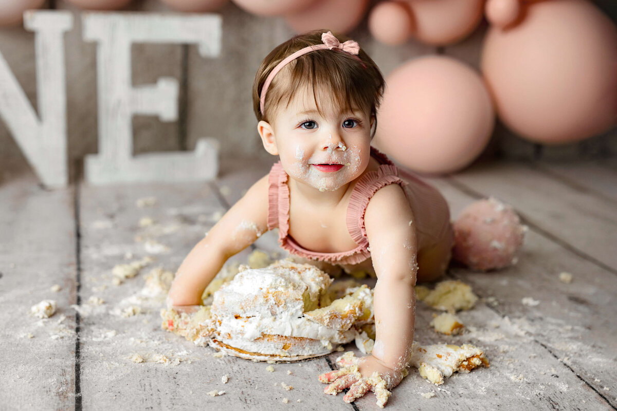 little girl in a pink outfit crawling on her birthday cake at her one year cake smash session with pink balloons