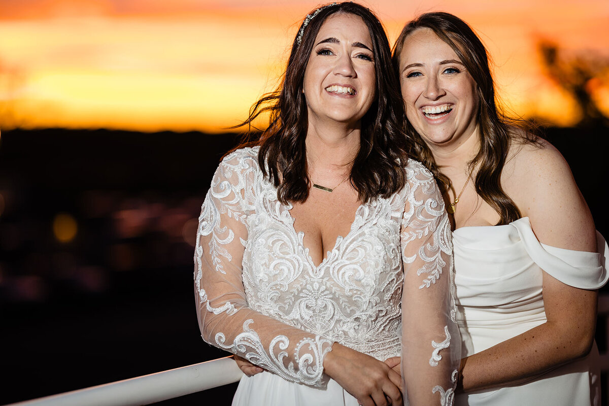 Two brides laughing together, with a beautiful sunset in the background, showcasing their joy and the romantic setting.