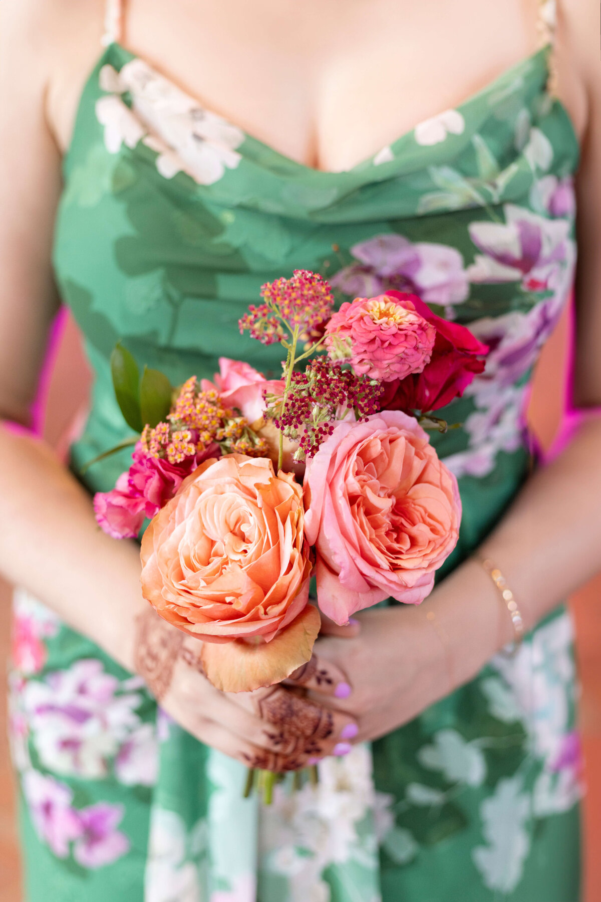 A woman in a floral green dress holds a bouquet of pink and orange roses mixed with small red flowers. Her nails are painted lavender, and she wears a bracelet. The background is softly blurred.