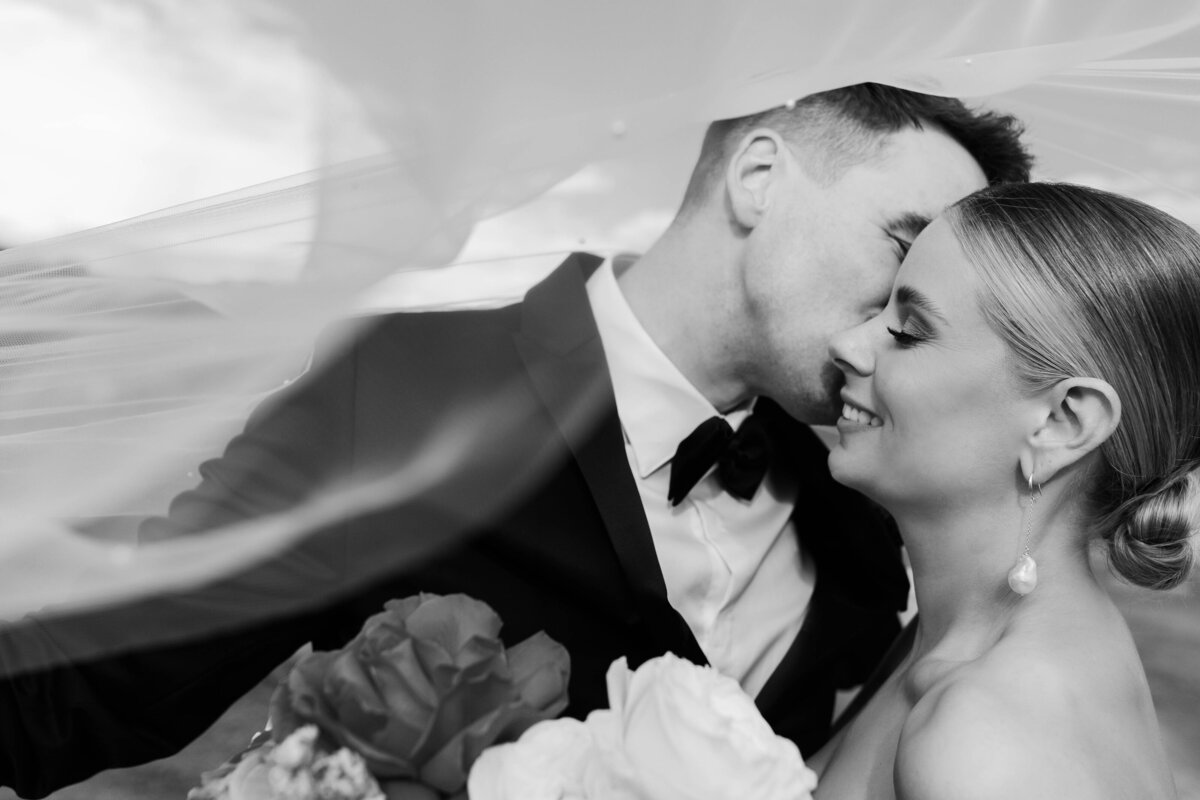 A black and white image of a couple on their wedding day. The groom is kissing the bride on the cheek. The bride holds a bouquet of flowers.