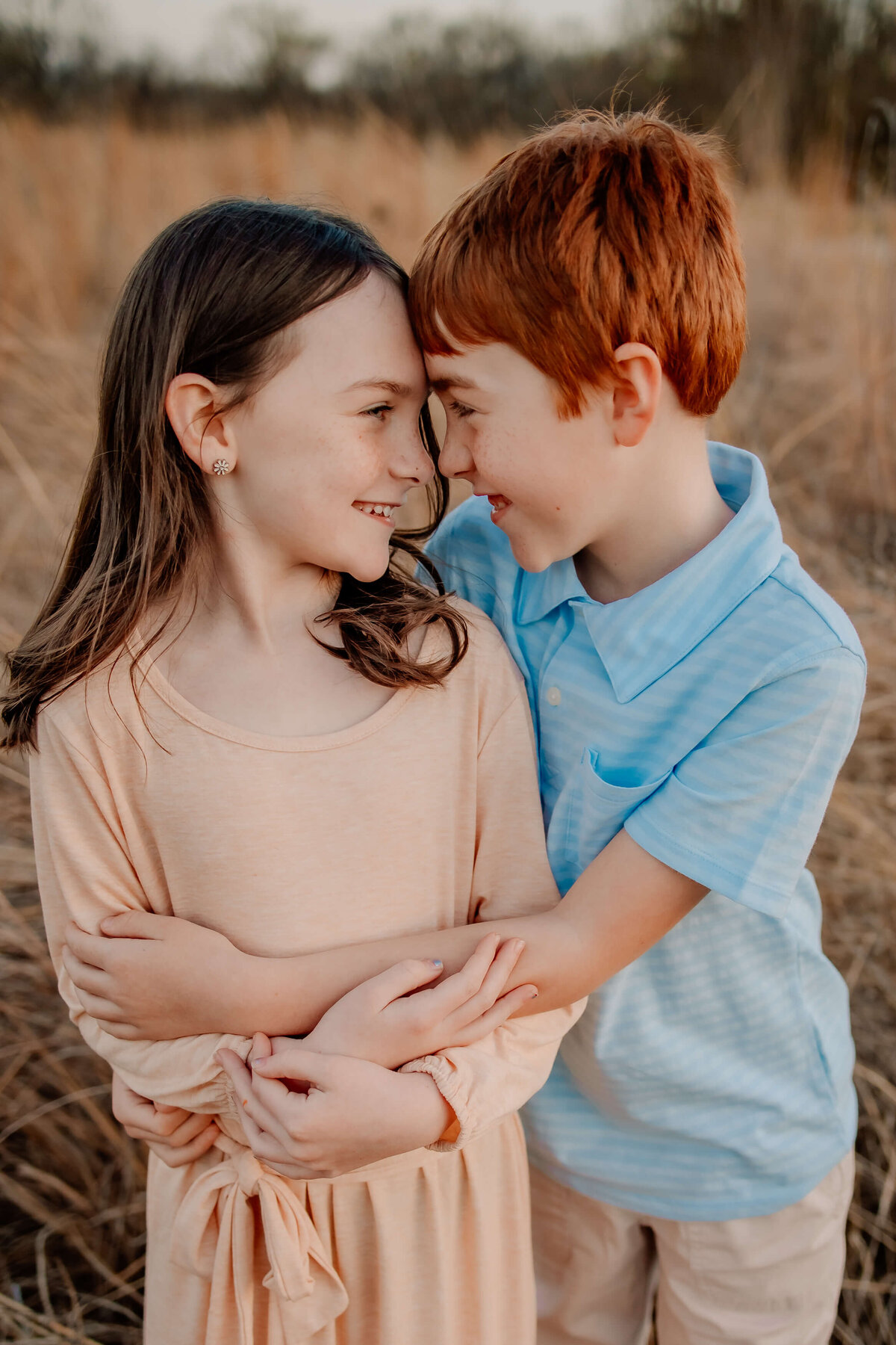 a red haired boy holding onto a brown hared girl both about 8 years old, standing in a field looking at eachother nose to nose