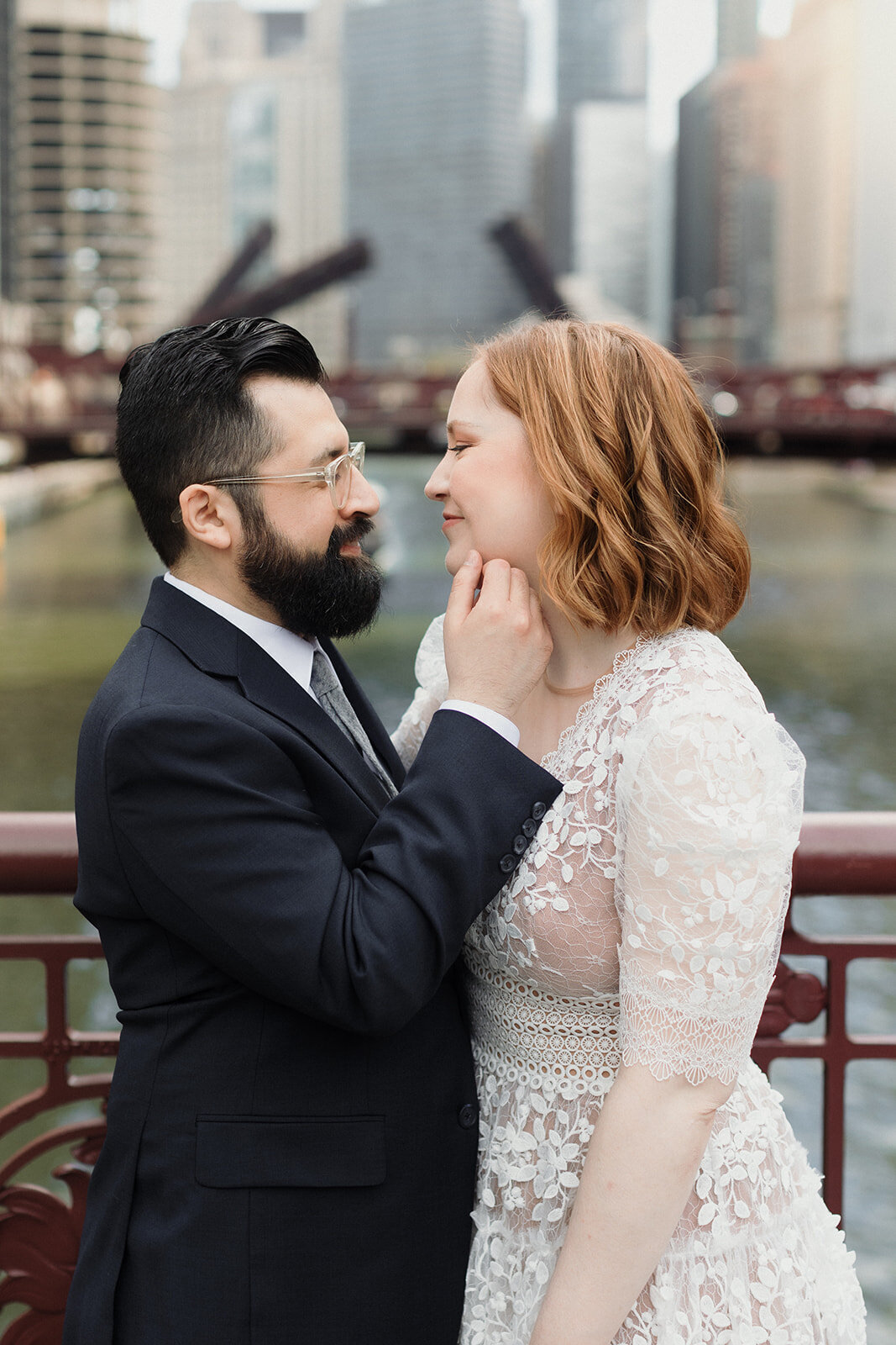Couple standing on Chicago River steel bridge. They're in wedding clothes and standing very close, groom has is hand on bride's face and he's about to kiss her.