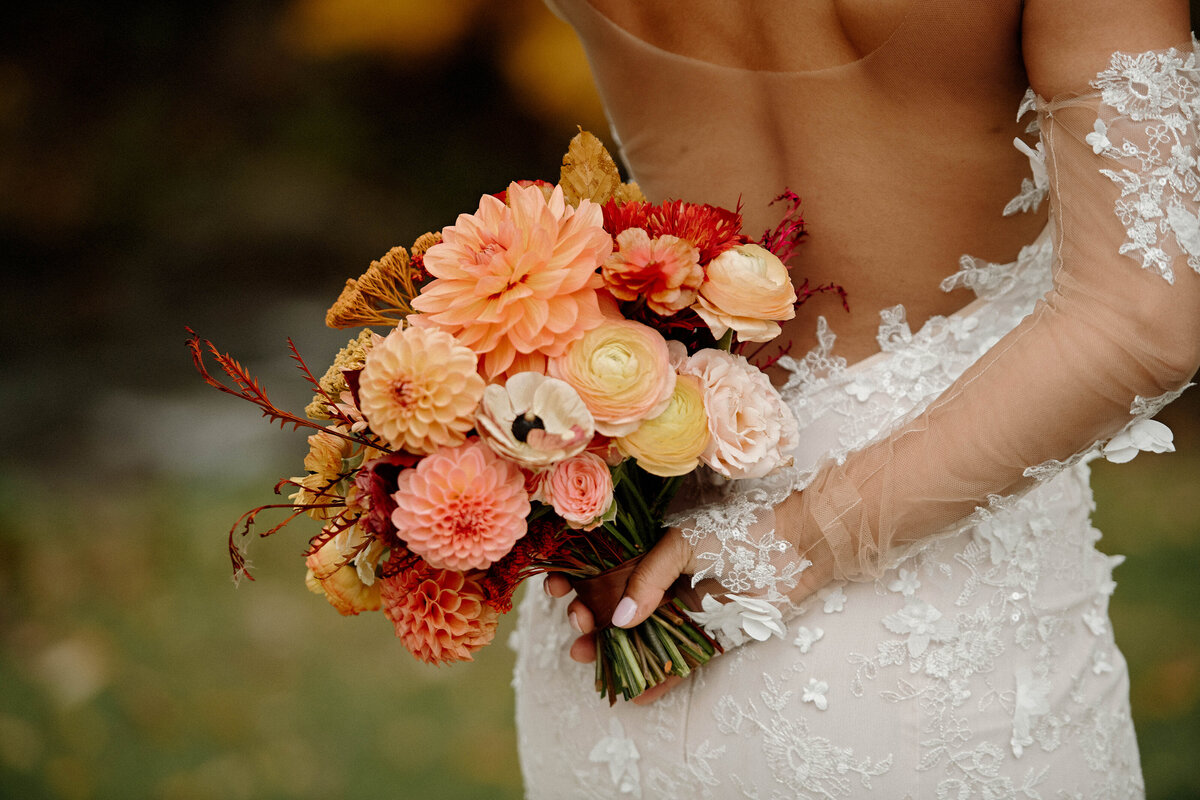 A Black bride in a beaded lace gown holding a bridal bouquet of dahlias and ranunculus behind her back