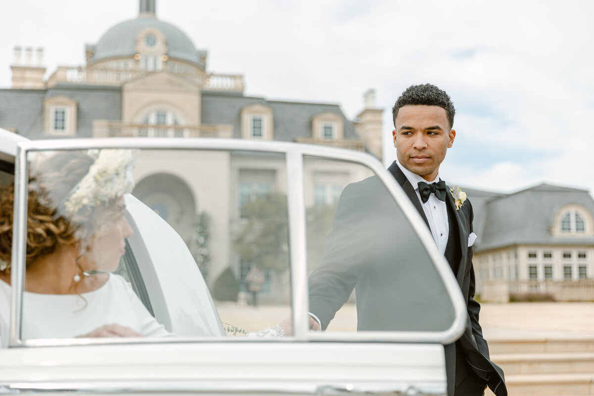 Handsome groom in tux helps his bride out of a white Rolls Royce outside of their chateau wedding venue