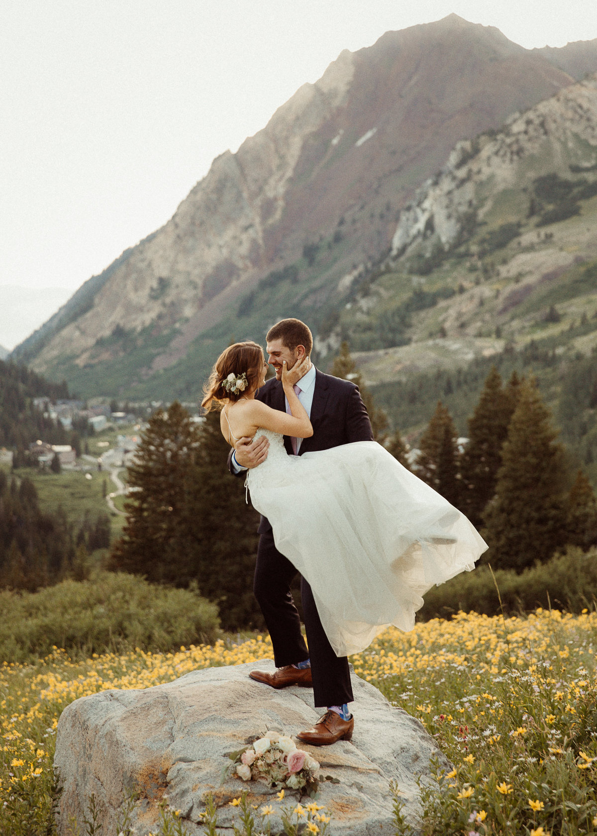 groom picks up bride while standing on a rock, surrounded by yellow flowers and Alta Mountains in the background