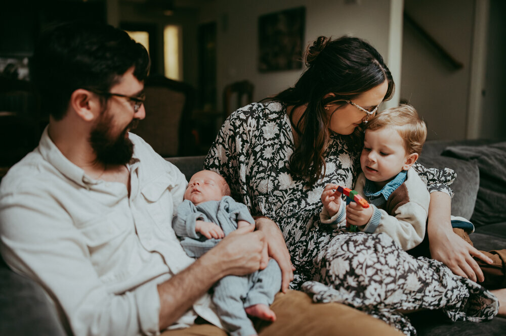 Candid newborn family photo—father holding sleeping baby while mother kisses toddler’s head, capturing love and everyday connection in a relaxed in-home session.