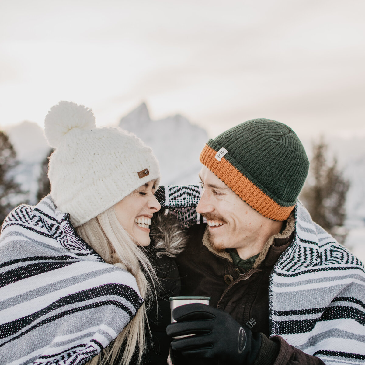 engaged couple laughing and  wrapped up in a blanket in front of the Tetons for their winter engagement session with jackson hole photographers