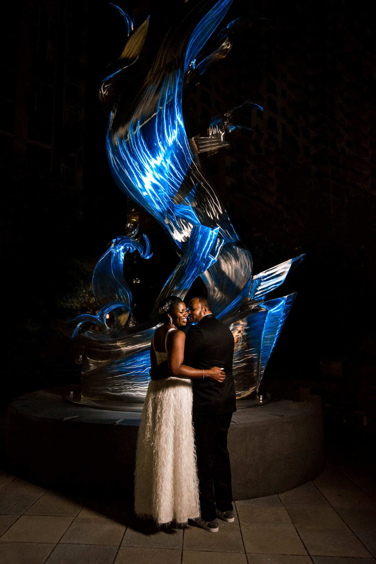 Nighttime-portrait-of-bride-and-groom-nuzzling-in-front-of-blue-lit-up-sculpture-in-Romare-Bearden-Park-in-Charlotte