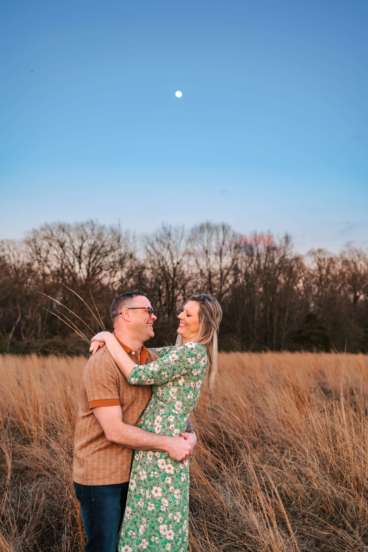 A man and woman are standing together in a field of tall grass  with their arms around each other smiling at each other and laughing. The moon is showing in the blue sky