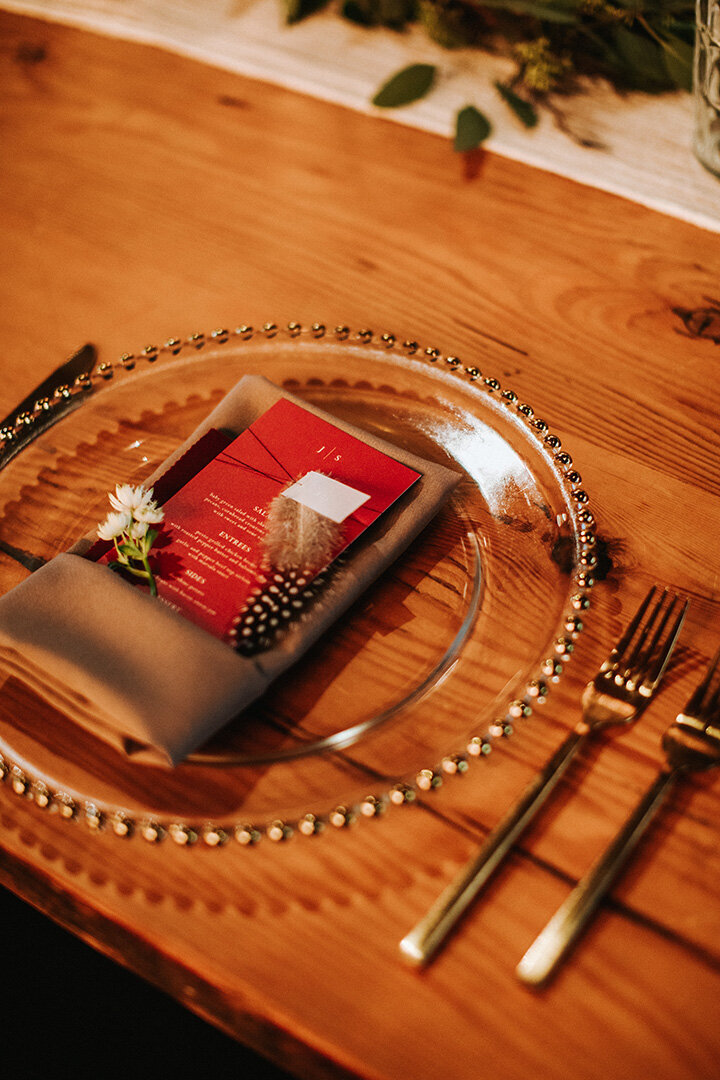 Red card stock dinner menu set atop a gold napkin with flowers on a clear plate and gold silverware.