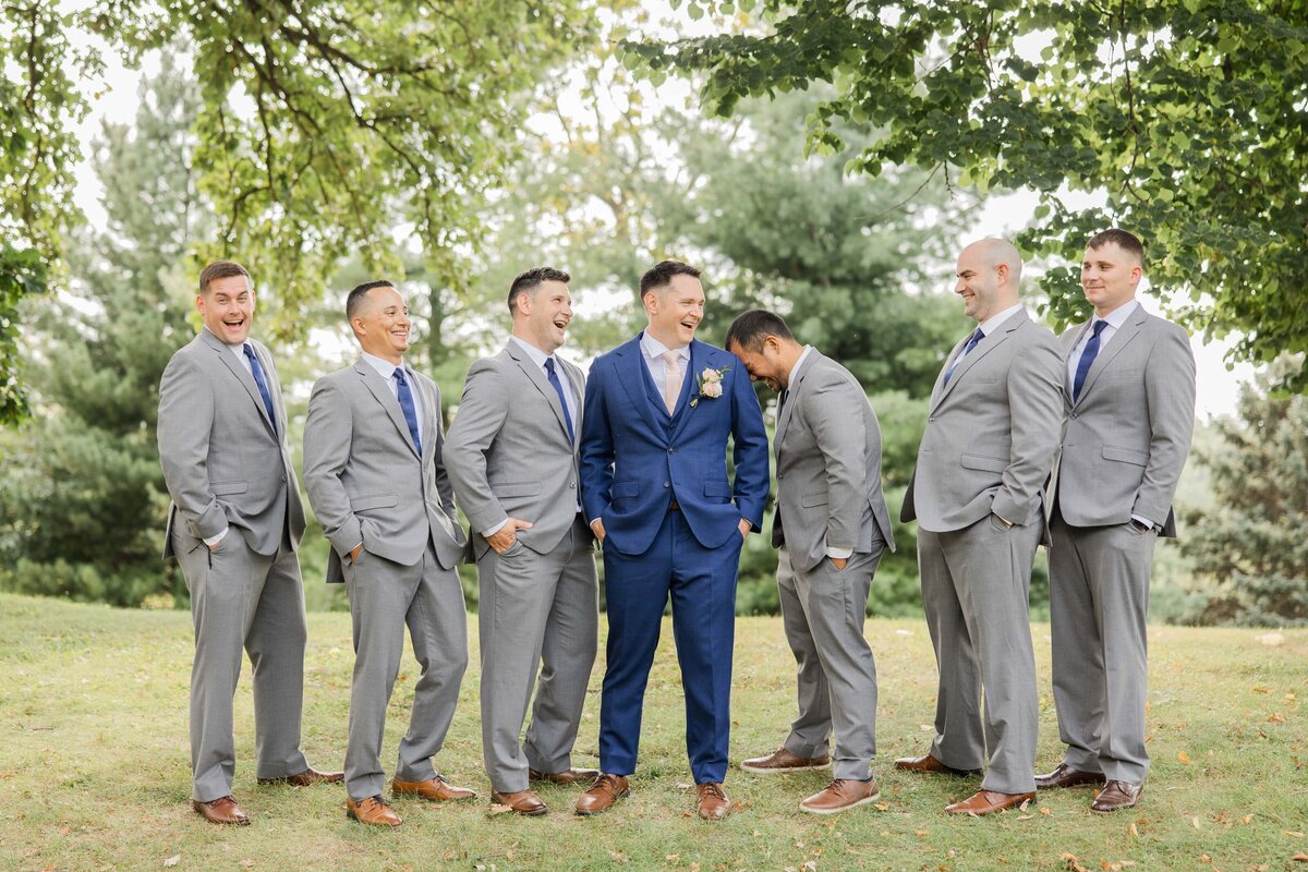 A group of seven men in suits, laughing together in a park during an Iowa wedding, with the groom in a blue suit, surrounded by groomsmen in gray.