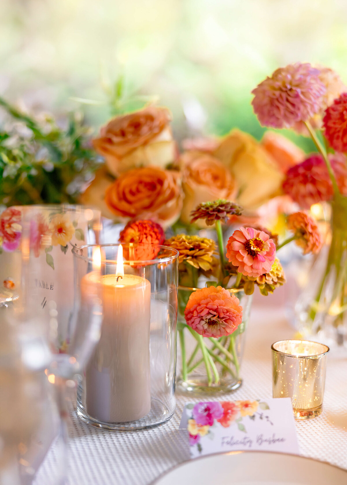 A beautifully arranged table centerpiece featuring vibrant orange and pink flowers in glass vases, surrounding a lit candle. The soft lighting and floral decor create an elegant and inviting atmosphere.